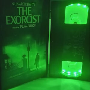 May include: A green-lit display featuring the movie title "The Exorcist" by William Peter Blatty and directed by William Friedkin. The display includes a DVD case and a glowing VHS tape replica, both illuminated with green lights.