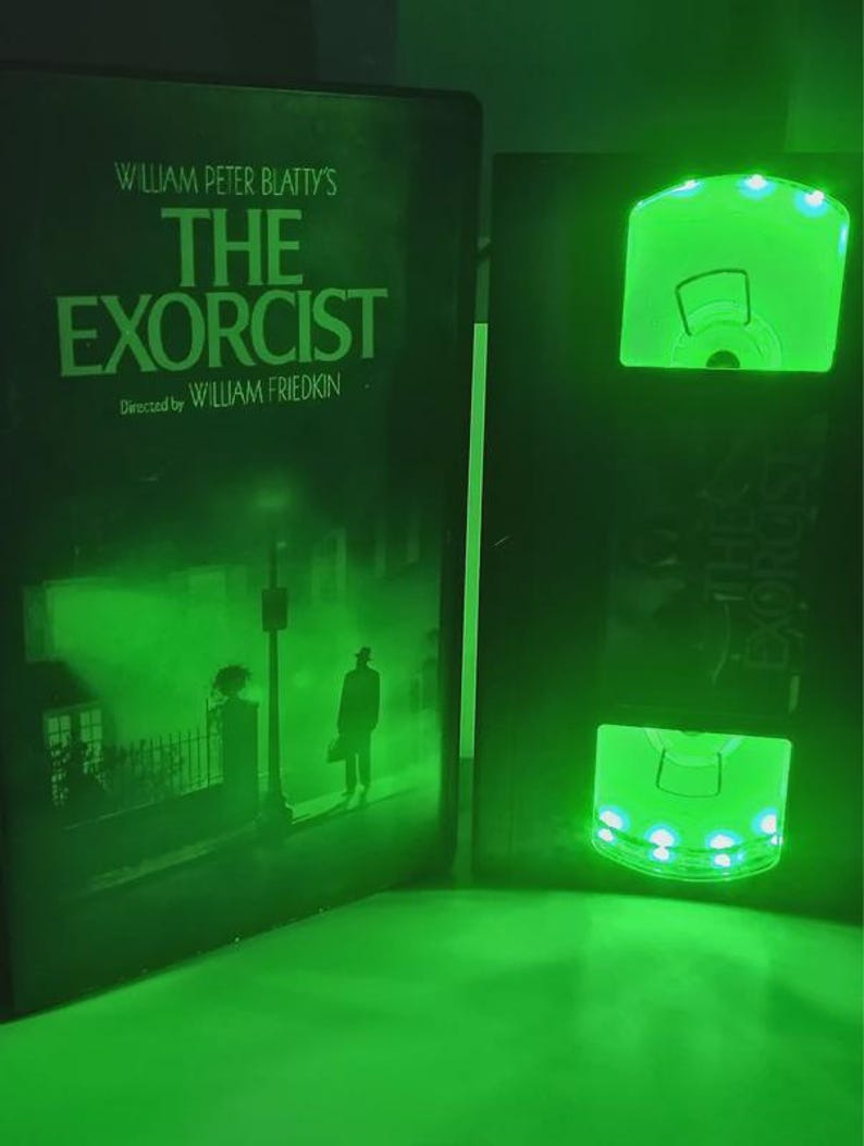 May include: A green-lit display featuring the movie title "The Exorcist" by William Peter Blatty and directed by William Friedkin. The display includes a DVD case and a glowing VHS tape replica, both illuminated with green lights.