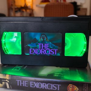 May include: A black VHS tape with the movie title "The Exorcist" and a green LED light. The tape is placed on top of a book with the same title. The image features a horror movie theme.