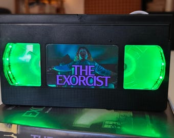 May include: A black VHS tape with the movie title "The Exorcist" and a green LED light. The tape is placed on top of a book with the same title. The image features a horror movie theme.