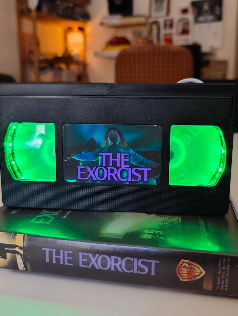 May include: A black VHS tape with the movie title "The Exorcist" and a green LED light. The tape is placed on top of a book with the same title. The image features a horror movie theme.