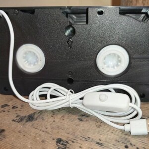 May include: A black VHS tape converted into a lamp. The lamp has two white spools, a white power cord, an on/off switch, and a USB connector. The lamp is sitting on a wooden surface.