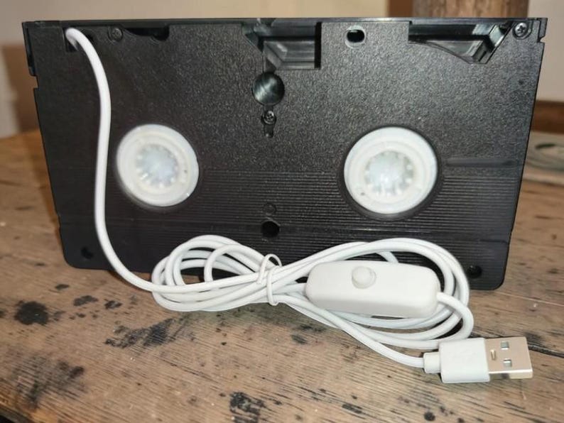 May include: A black VHS tape converted into a lamp. The lamp has two white spools, a white power cord, an on/off switch, and a USB connector. The lamp is sitting on a wooden surface.