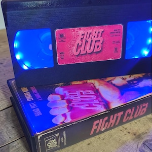 Può includere: Una cassetta VHS nera con un'etichetta Fight Club e luci LED blu brillante che illuminano le bobine interne, posizionata sopra la custodia originale che mostra un pugno e il titolo Fight Club in grassetto rosa.
