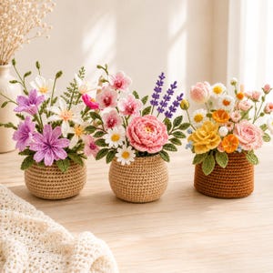 May include: Three crocheted flower arrangements in round, textured pots. The left pot holds purple and white flowers. The center pot features pink orchids, a large pink rose, and daisies. The right pot contains yellow, pink, and white flowers.