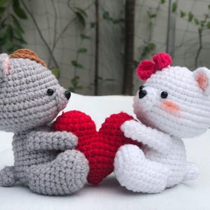 Peut inclure: Deux animaux en peluche crochetés, l'un gris et l'autre blanc, tenant un cœur rouge crocheté. L'animal gris a des oreilles brunes et l'animal blanc a un nœud rouge.