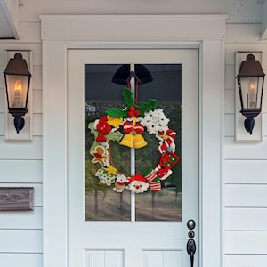 Puede incluir: Una corona navide&ntilde;a circular con varios adornos de ganchillo que incluyen una cara de Pap&aacute; Noel, un hombre de jengibre, un bast&oacute;n de caramelo, campanas, hojas de acebo, un copo de nieve y calcetines unidos a un aro de madera.
