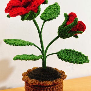 May include: A crocheted plant with red flowers and green leaves in a brown pot. The plant is made of yarn and is a handmade craft.