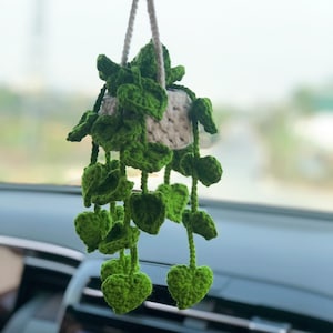 Puede incluir: Una planta colgante de ganchillo con hojas verdes y una maceta blanca. La planta está colgada del espejo retrovisor de un coche.