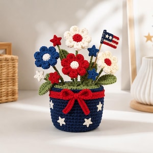 May include: A crocheted arrangement featuring red, white, and blue flowers and stars in a dark blue pot. The pot is decorated with white stars and a red bow. A small American flag on a wooden stick is tucked among the crocheted green leaves.