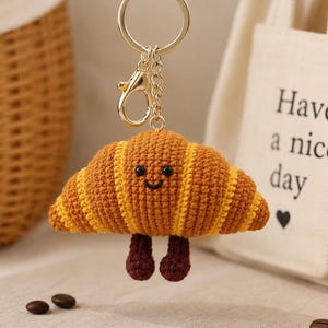 May include: A gold-toned metal keychain featuring a crocheted croissant charm. The croissant is golden-brown with yellow stripes, small black bead eyes, a stitched smile, and two dangling dark brown crocheted legs attached to the bottom.