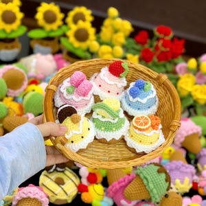 May include: A wicker basket displays several crocheted miniature cakes, each with unique toppings such as strawberries, blueberries, and citrus slices. The cakes are in various colours, including pink, yellow, and blue. The background features crocheted flowers and other food items.