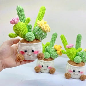Puede incluir: Tres cactus de ganchillo hechos a mano en macetas blancas con caras sonrientes, tierra marrón y cactus verdes con flores rosas y amarillas. Las macetas tienen pies marrones.