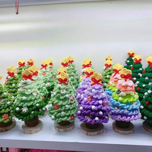 以下が含まれることがあります： 緑、紫、虹色など、様々な色の手編みクリスマスツリーのコレクション。各ツリーは黄色の星、赤いリボン、白いポンポンで飾られ、木の台座に置かれています。お祝いのホリデーデコレーション。