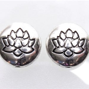 Könnte beinhalten: Zwei silberfarbene, runde Perlen mit einem Lotusblumen-Design. Die Lotusblume ist auf der Oberfläche jeder Perle geprägt. Die Perlen werden wahrscheinlich für die Schmuckherstellung oder andere Bastelprojekte verwendet.