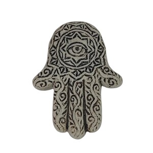 Op de afbeelding: Een witte keramische hamsa-hand met een zwart oog en ingewikkelde ontwerpen. De hamsa is een symbool van bescherming en geluk.