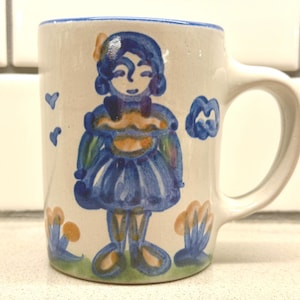 May include: A ceramic mug with a cream-colored base featuring a hand-painted blue illustration of a figure wearing a dress. The design includes small blue hearts, a cloud, and stylized floral elements in blue and tan at the base of the mug.