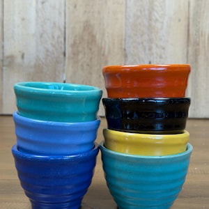 May include: A set of six small ceramic bowls stacked in two columns. The left stack features a teal bowl, a light blue bowl, and a dark blue bowl. The right stack features an orange bowl, a black bowl, a yellow bowl, and a teal bowl at the base.