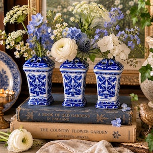 Può includere: Tre vasi in porcellana blu e bianca con composizioni floreali poggiano su libri antichi. I vasi hanno una forma quadrata con un motivo floreale blu. I fiori sono bianchi e blu, creando un'esposizione classica ed elegante.