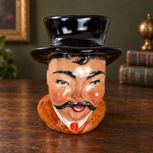 May include: A ceramic character jug shaped as a person wearing a glossy black top hat. The figure features painted blue eyes, dark eyebrows, a prominent black moustache, rosy cheeks, and a brown patterned coat with a small red necktie detail.