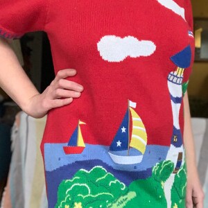 Puede incluir: Su&eacute;ter de punto rojo con tem&aacute;tica n&aacute;utica. El su&eacute;ter presenta un faro, dos veleros, una nube y una isla verde con flores amarillas.