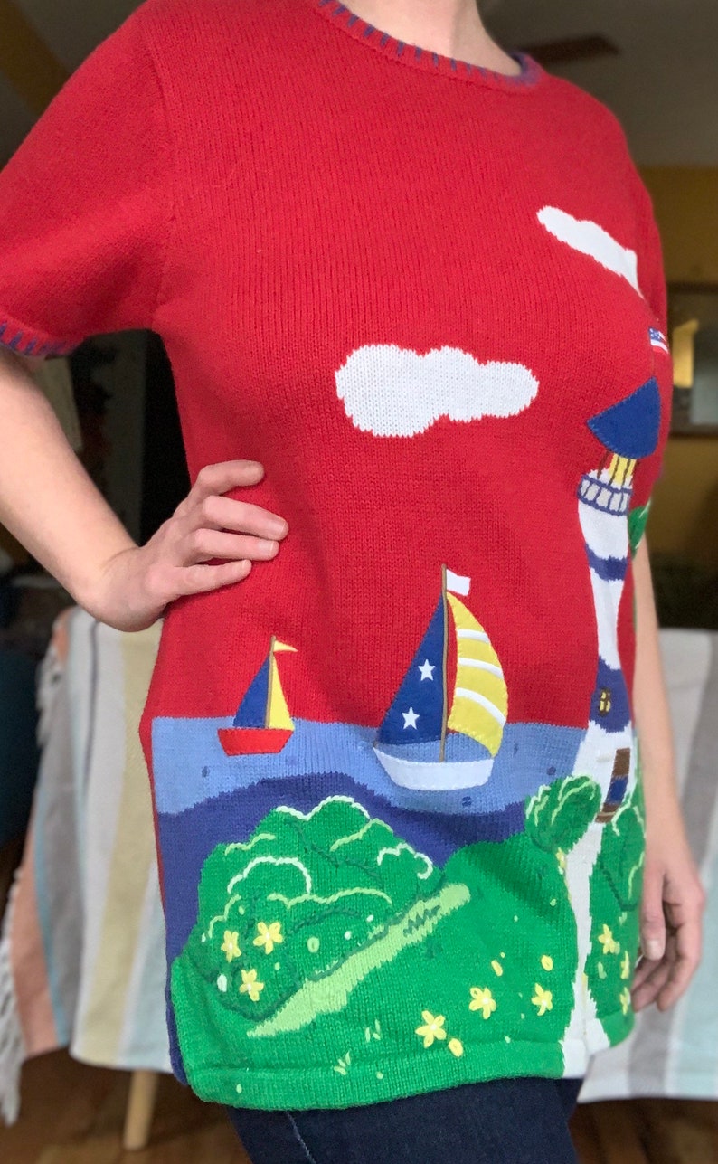 Puede incluir: Su&eacute;ter de punto rojo con tem&aacute;tica n&aacute;utica. El su&eacute;ter presenta un faro, dos veleros, una nube y una isla verde con flores amarillas.