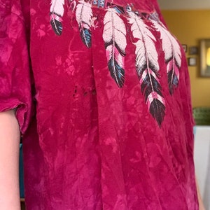 May include: A red tie-dye t-shirt with a graphic of a Native American inspired design featuring feathers and geometric patterns.
