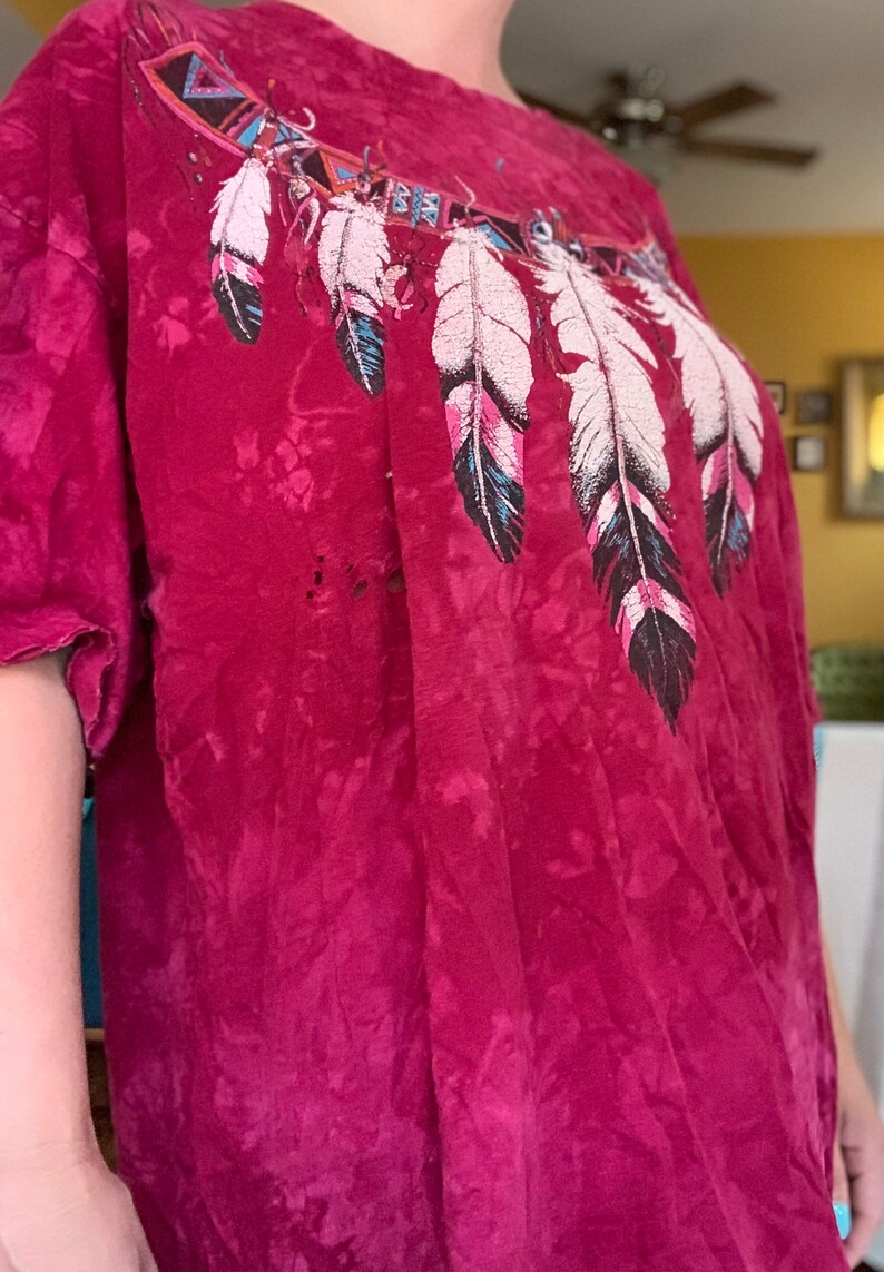 May include: A red tie-dye t-shirt with a graphic of a Native American inspired design featuring feathers and geometric patterns.