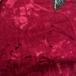 May include: A close-up of a maroon fabric with several small holes. The fabric has a faded, tie-dye pattern.