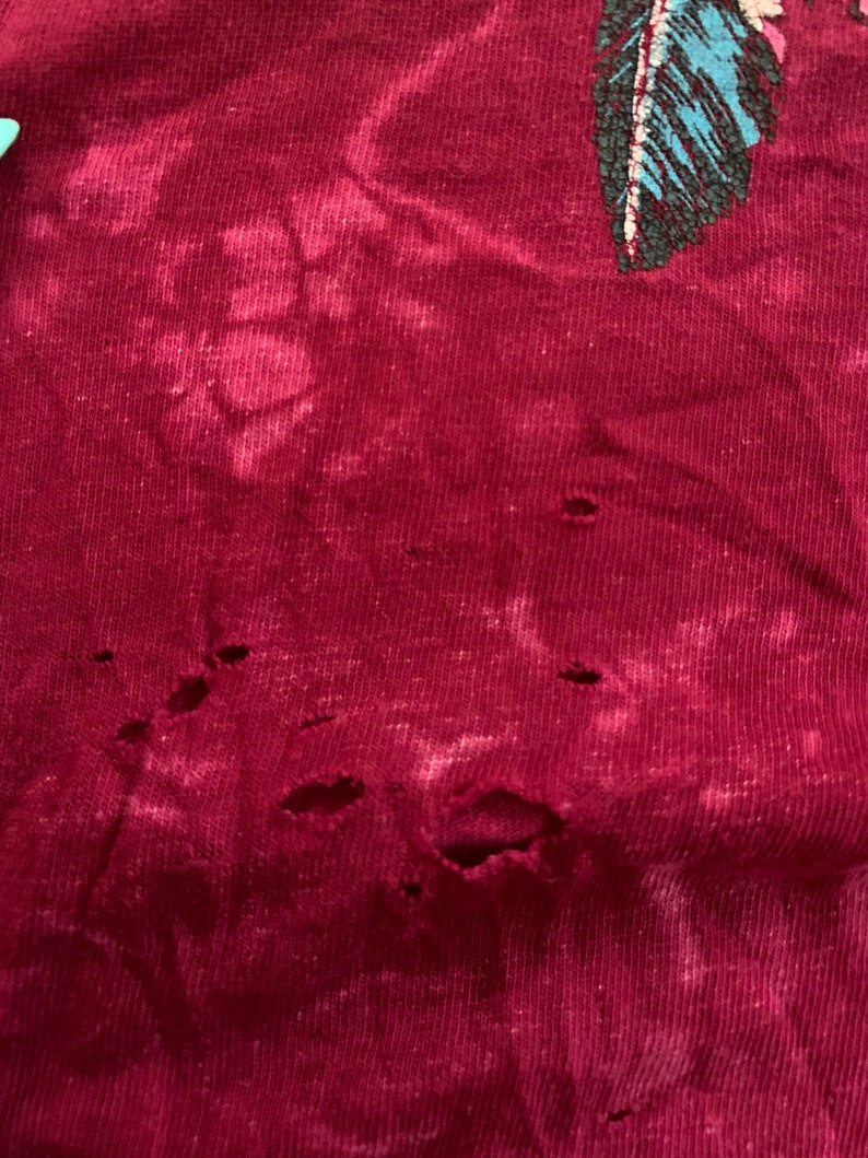 May include: A close-up of a maroon fabric with several small holes. The fabric has a faded, tie-dye pattern.