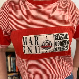 May include: A short-sleeved t-shirt with red and white horizontal stripes. A large rectangular patch on the chest features the word MARINE, a compass rose graphic with a ship, and a grid of numbers including 21, 05, 11, 09, 88, 16, 07, 12, 06.