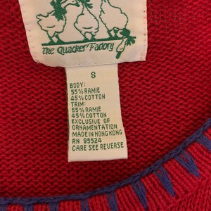 Puede incluir: Un su&eacute;ter rojo con ribetes azules y una etiqueta blanca que dice "the Quacker Factory" y tiene una talla "S". La etiqueta tambi&eacute;n enumera la composici&oacute;n de la tela como 55% ramie y 45% algod&oacute;n para el cuerpo y el ribete. La etiqueta tambi&eacute;n dice "Exclusive of ornamentation, Made in Hong Kong, RN 95524, Care see reverse."