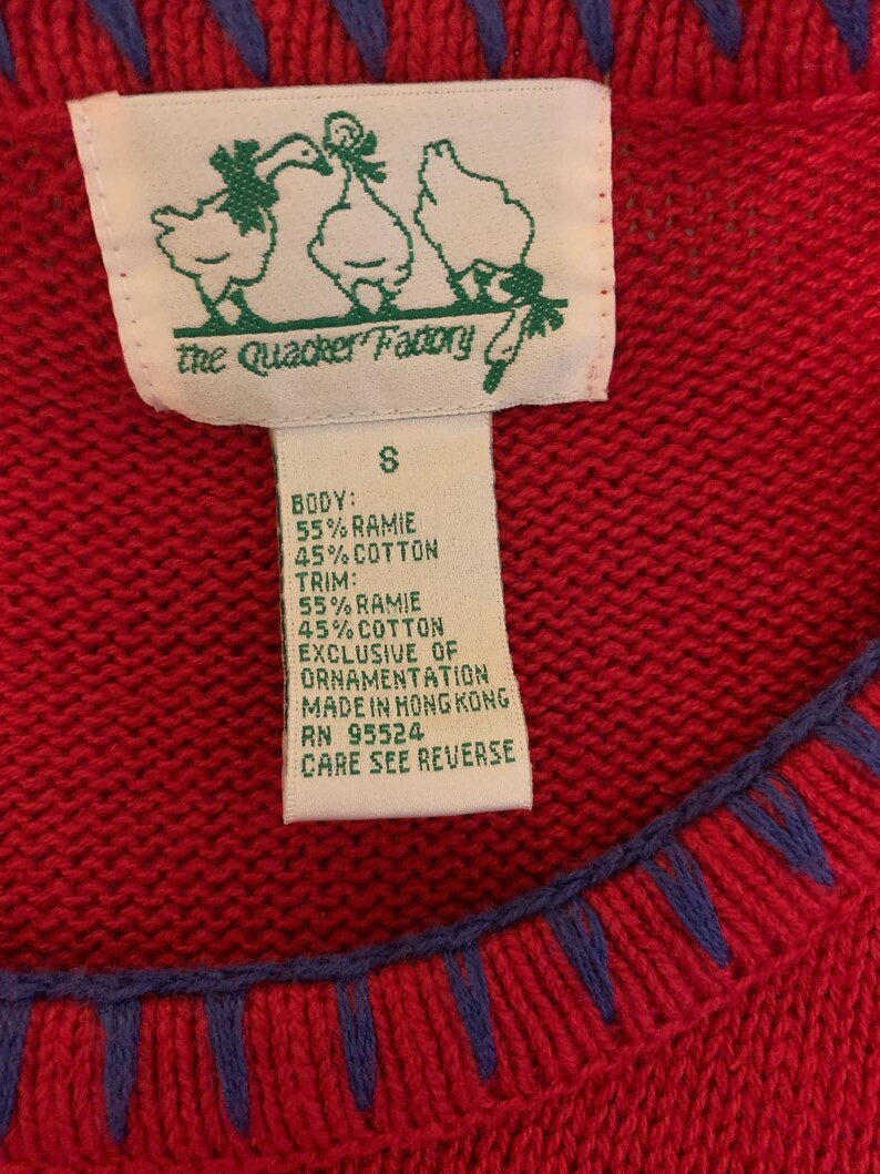 Puede incluir: Un su&eacute;ter rojo con ribetes azules y una etiqueta blanca que dice "the Quacker Factory" y tiene una talla "S". La etiqueta tambi&eacute;n enumera la composici&oacute;n de la tela como 55% ramie y 45% algod&oacute;n para el cuerpo y el ribete. La etiqueta tambi&eacute;n dice "Exclusive of ornamentation, Made in Hong Kong, RN 95524, Care see reverse."