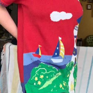 Puede incluir: Su&eacute;ter de punto rojo con tem&aacute;tica n&aacute;utica. El su&eacute;ter presenta un faro, un velero y nubes en blanco, azul y amarillo.