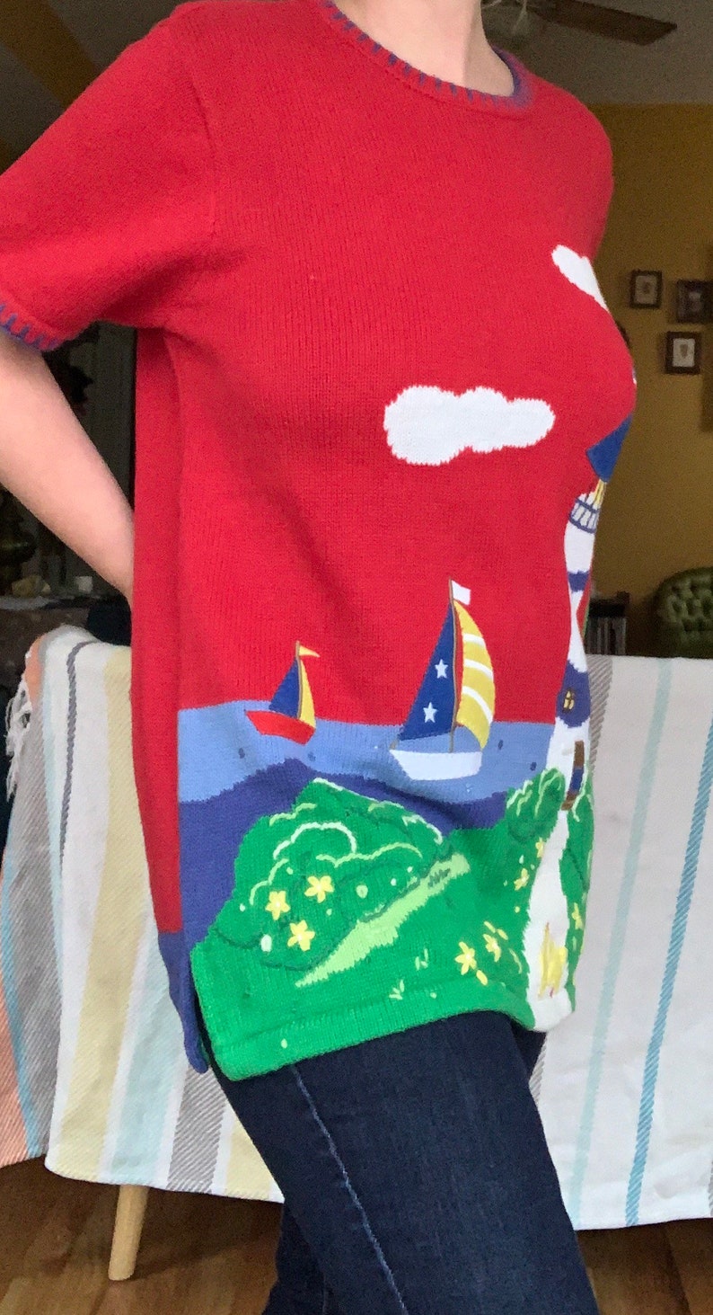 Puede incluir: Su&eacute;ter de punto rojo con tem&aacute;tica n&aacute;utica. El su&eacute;ter presenta un faro, un velero y nubes en blanco, azul y amarillo.