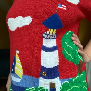 Puede incluir: Su&eacute;ter de punto rojo con un faro blanco y azul, un velero y &aacute;rboles verdes. El faro tiene un techo azul y una peque&ntilde;a bandera estadounidense en la parte superior.