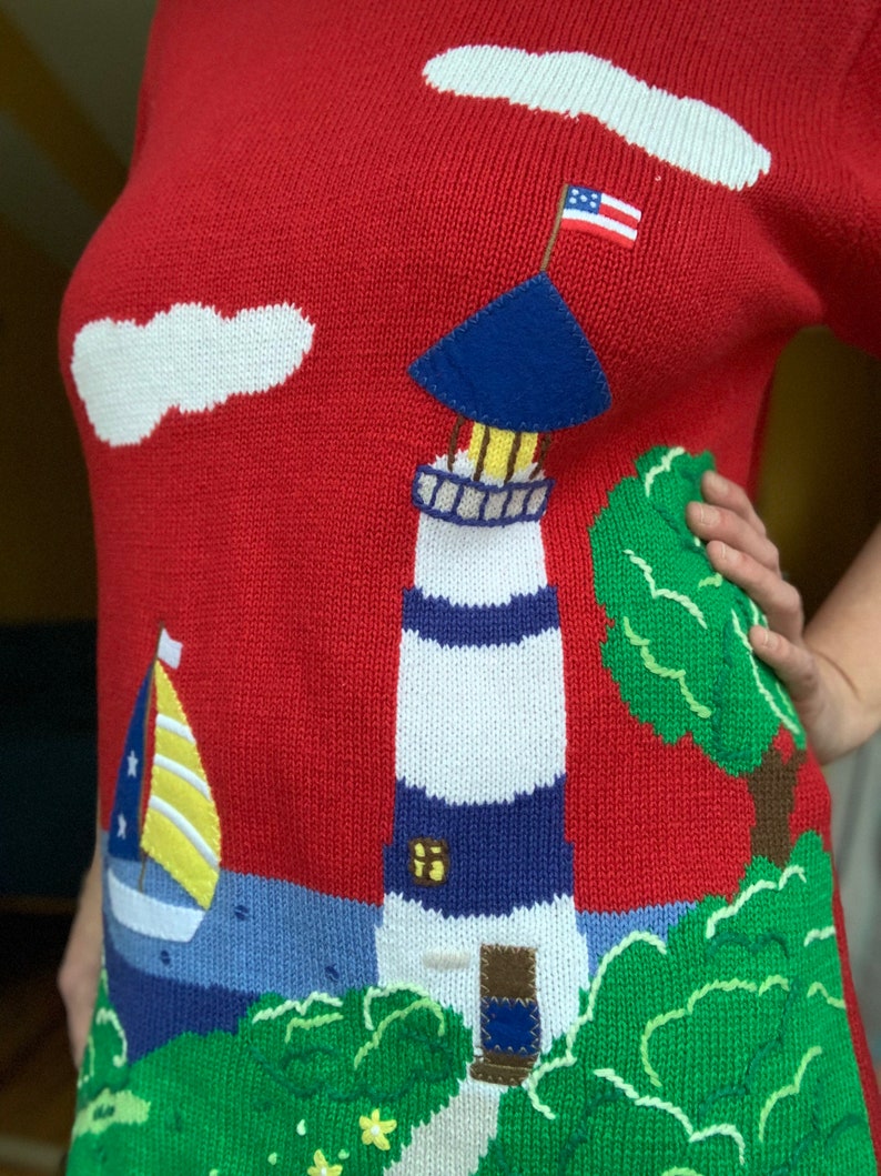 Puede incluir: Su&eacute;ter de punto rojo con un faro blanco y azul, un velero y &aacute;rboles verdes. El faro tiene un techo azul y una peque&ntilde;a bandera estadounidense en la parte superior.