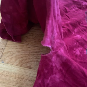 May include: A close-up of a red t-shirt with a faded tie-dye pattern. The shirt has a small tear near the shoulder seam.