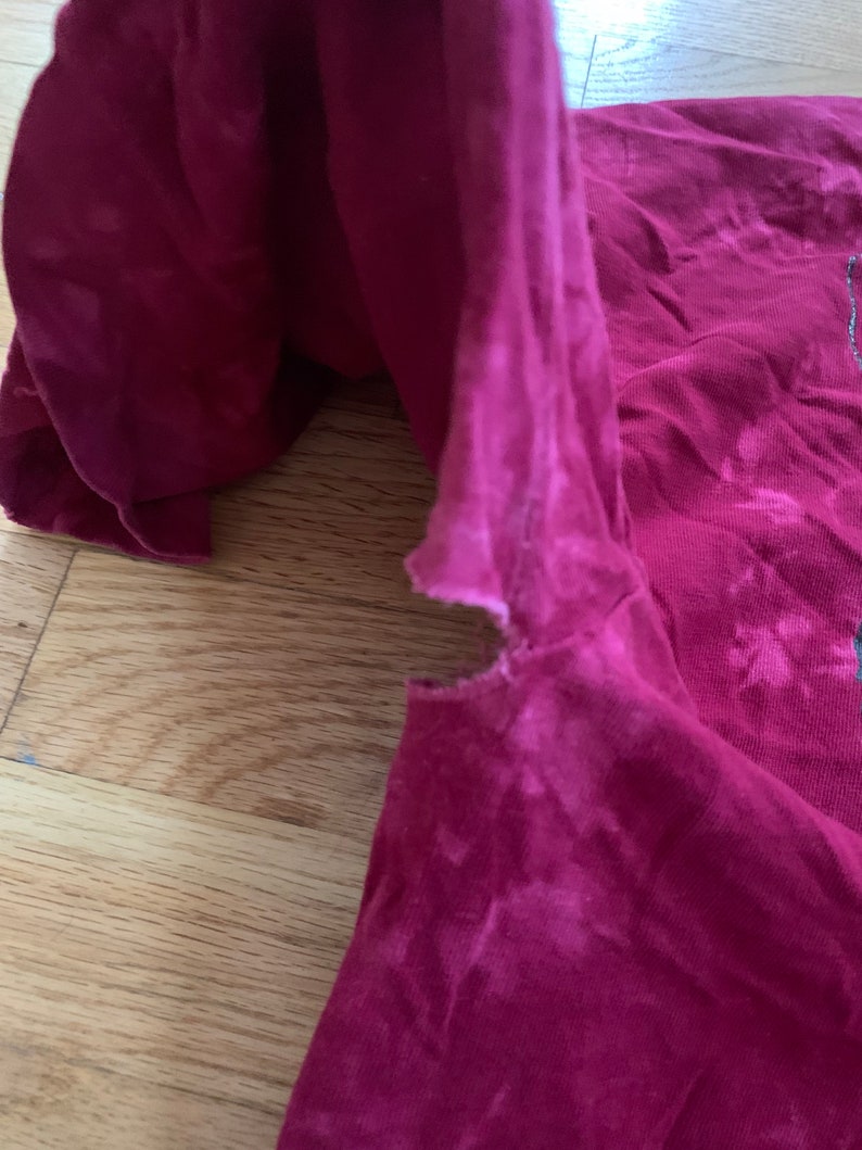 May include: A close-up of a red t-shirt with a faded tie-dye pattern. The shirt has a small tear near the shoulder seam.