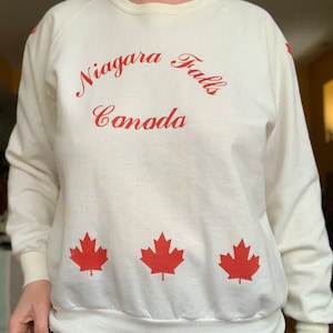 May include: White sweatshirt with red lettering that says "Niagara Falls Canada" and three red maple leaf designs.