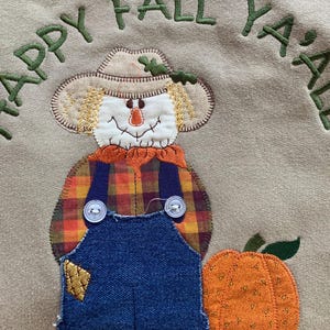 May include: Tan sweatshirt with an embroidered scarecrow and pumpkin design. The scarecrow wears a straw hat, plaid shirt, and denim overalls. The words "HAPPY FALL YA'LL" are embroidered in green.