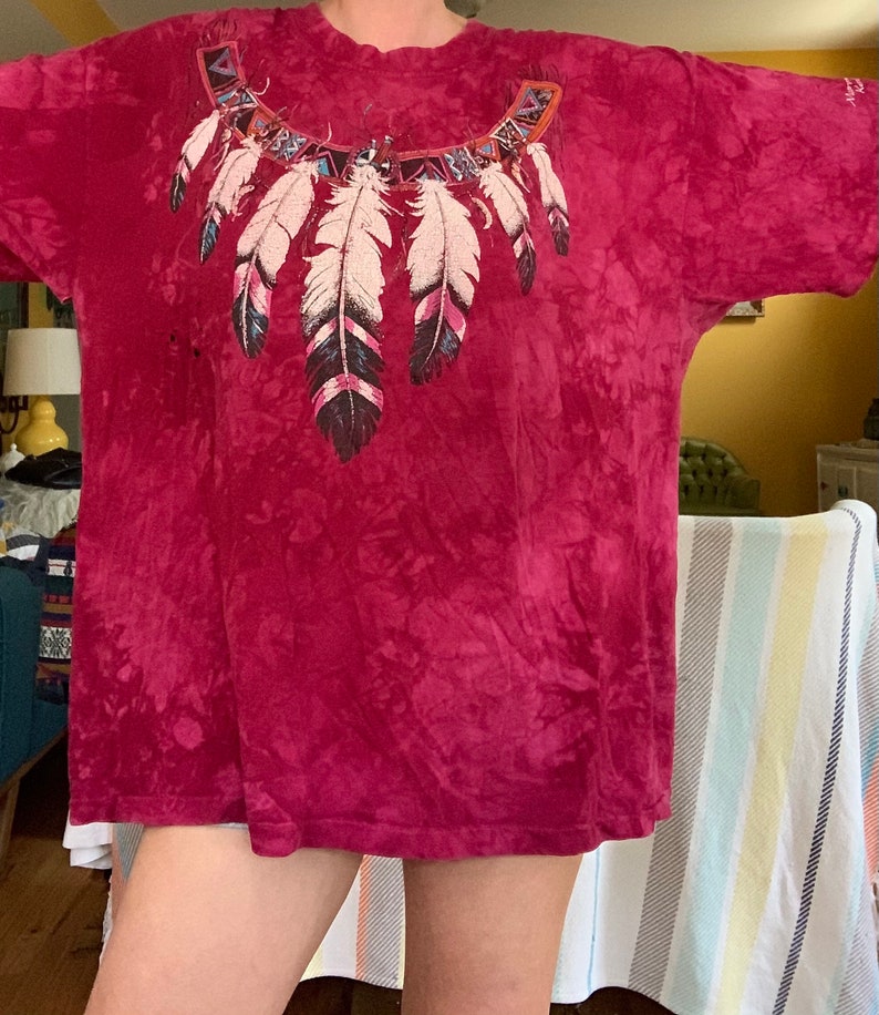 May include: A red tie-dye t-shirt with a colorful Native American inspired feather design on the front.