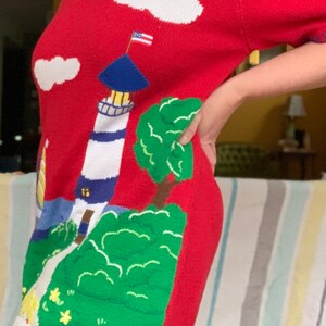 Puede incluir: Su&eacute;ter de punto rojo con un dise&ntilde;o de faro, nubes y &aacute;rbol. El faro tiene un techo azul y rayas blancas y azules. El &aacute;rbol es verde y las nubes son blancas.