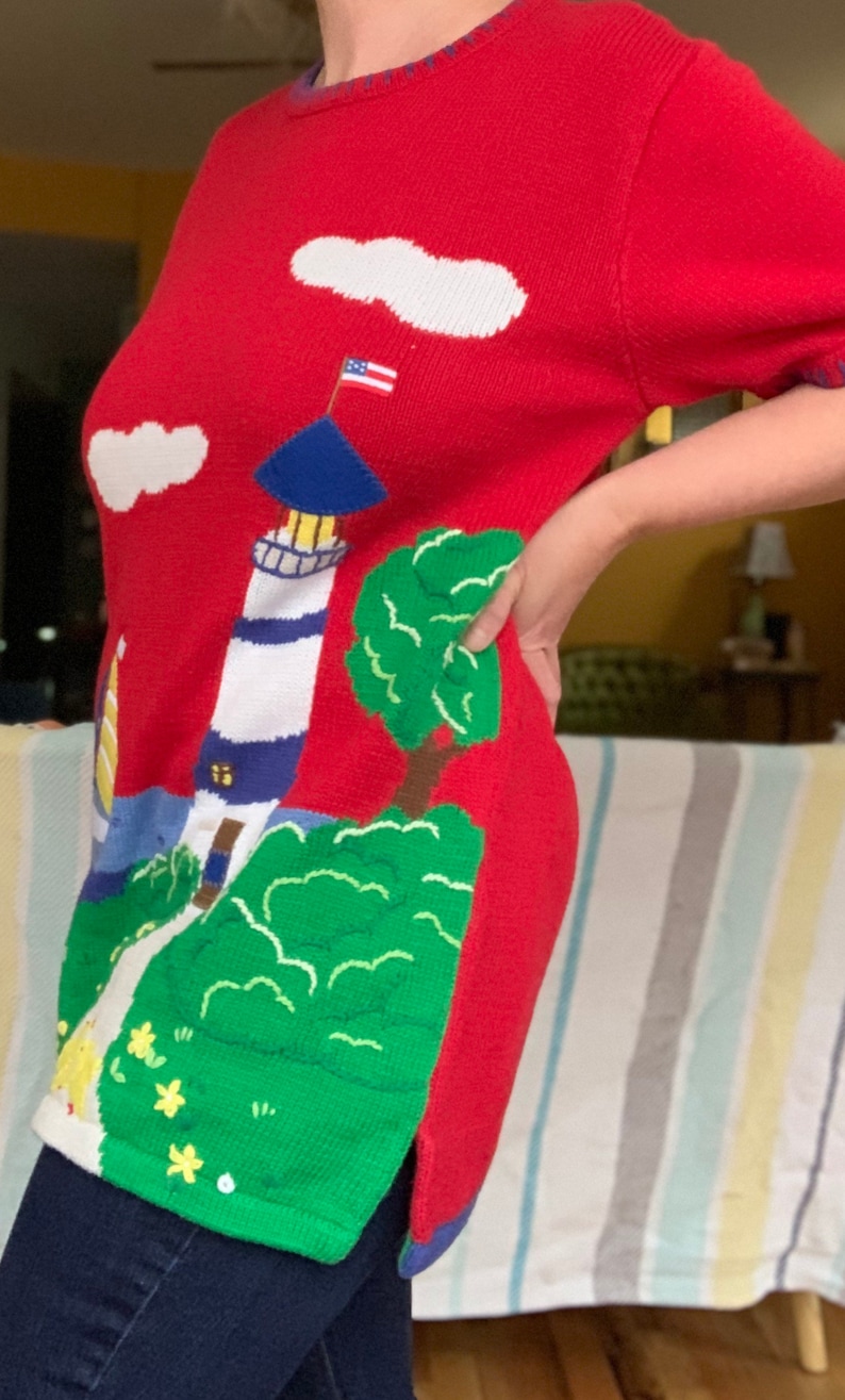 Puede incluir: Su&eacute;ter de punto rojo con un dise&ntilde;o de faro, nubes y &aacute;rbol. El faro tiene un techo azul y rayas blancas y azules. El &aacute;rbol es verde y las nubes son blancas.