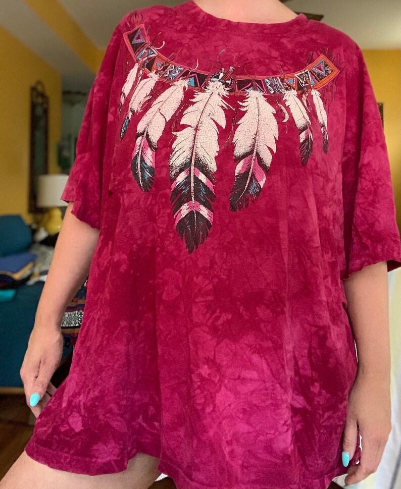 May include: A red tie-dye t-shirt with a Native American inspired design featuring feathers and geometric patterns.