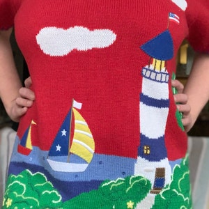 Puede incluir: Su&eacute;ter de punto rojo con un faro, un velero y nubes sobre un fondo azul y verde. El faro tiene un techo azul y rayas blancas. El velero tiene una vela amarilla con estrellas blancas. Las nubes son blancas. El fondo es azul y verde con un camino que lleva al faro.