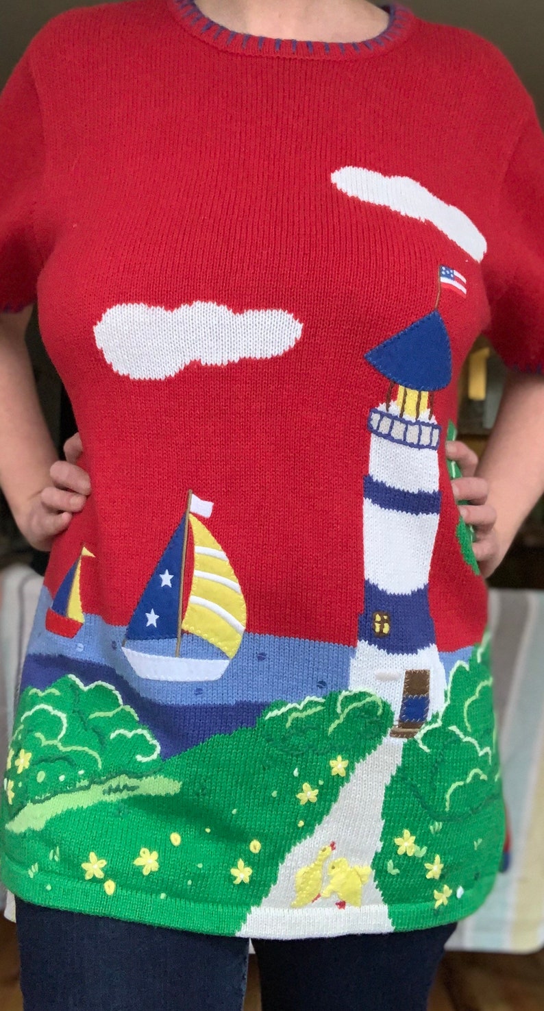 Puede incluir: Su&eacute;ter de punto rojo con un faro, un velero y nubes sobre un fondo azul y verde. El faro tiene un techo azul y rayas blancas. El velero tiene una vela amarilla con estrellas blancas. Las nubes son blancas. El fondo es azul y verde con un camino que lleva al faro.