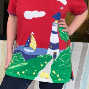 May include: Red knit sweater with a lighthouse, sailboat, clouds, and a small American flag. The sweater has a green, blue, and white landscape design.