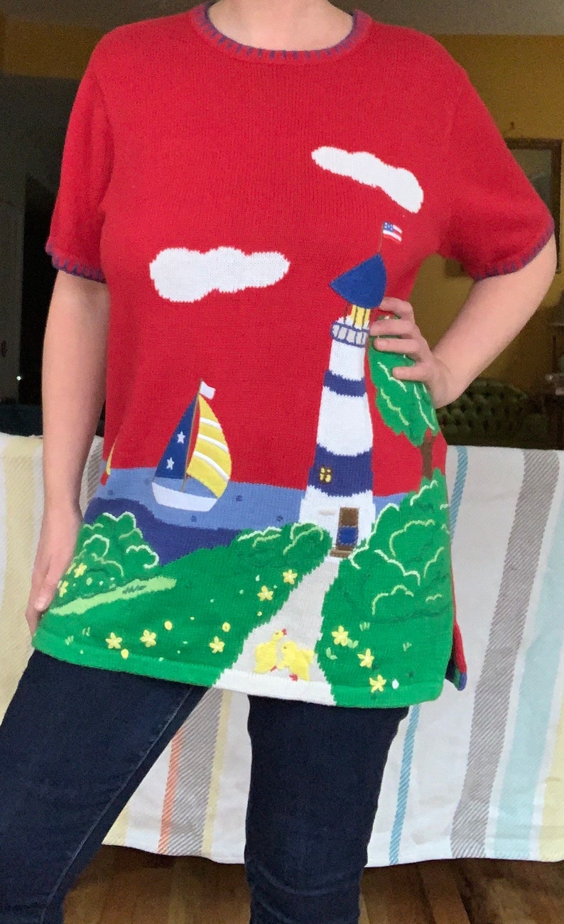 Puede incluir: Su&eacute;ter de punto rojo con un faro, un velero, nubes y una peque&ntilde;a bandera estadounidense. El su&eacute;ter tiene un dise&ntilde;o de paisaje verde, azul y blanco.