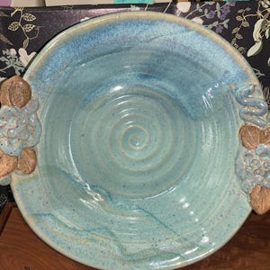 May include: A round ceramic bowl with a light blue glazed finish and a spiral pattern in the center. The bowl features two decorative handles on opposite sides, each adorned with sculpted blue flowers and brown leaves.